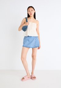 White lace strapless top, blue denim mini skirt, pink slide sandals, carrying a blue fabric bag, standing against a neutral background.