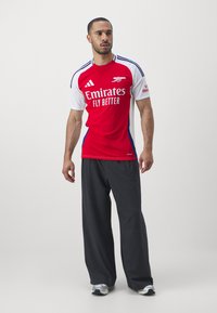 Tricou de fotbal Arsenal roșu și alb, cu logo și textul "Fly Better", asociat cu pantaloni gri largi și adidași.