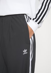 Black athletic sweatpants with elastic waistband, white side stripes, and a small silver Adidas logo on the upper left side.