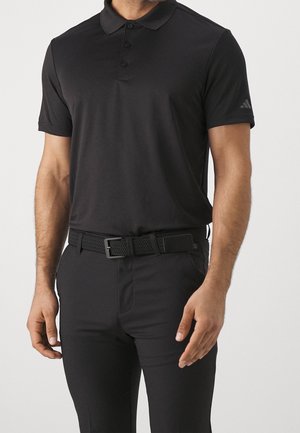 Homme portant un polo noir à manches courtes et un pantalon noir avec une ceinture tissée noire, debout les bras détendus le long du corps.