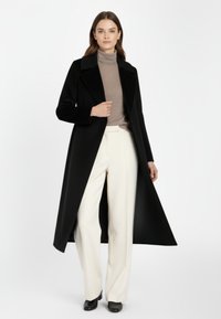 Zwarte wollen jas met een lange silhouet, brede revers en zijsplitten. Draagt over een beige turtleneck en witte wijde broek.