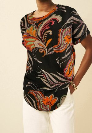 Czarna bluzka z krótkim rękawem z jaskrawymi, pomarańczowymi, zielonymi i różowymi kwiatowymi i spiralnymi wzorami, noszona z białymi spodniami i złotą bransoletką.