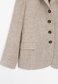Blazer en lin beige avec quatre boutons foncés sur le devant, une poche à rabat sur le côté gauche et des manches longues.