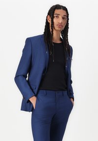 Costume bleu avec un blazer et un pantalon assortis, porté sur une chemise noire. Il présente un tissu lisse et des lignes épurées, avec des poches et des détails de boutons.