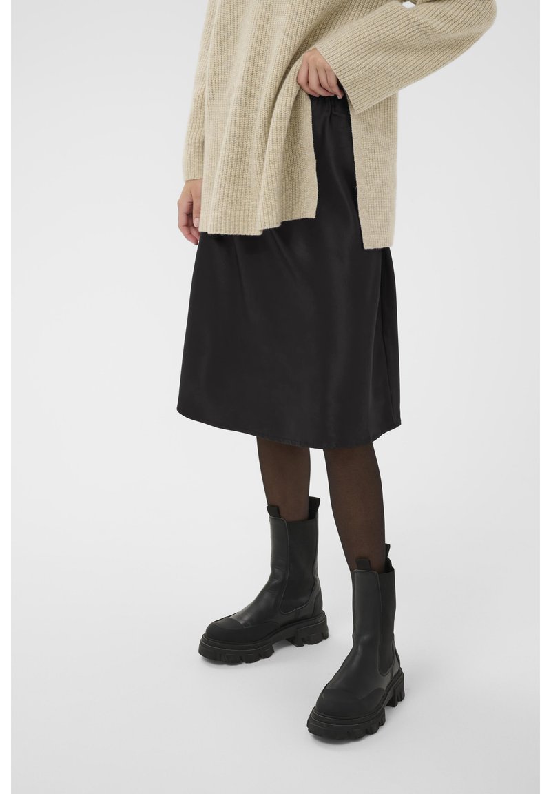 Jupe en satin noir associée à un pull côtelé crème et des bottines noires épaisses, sur fond blanc.