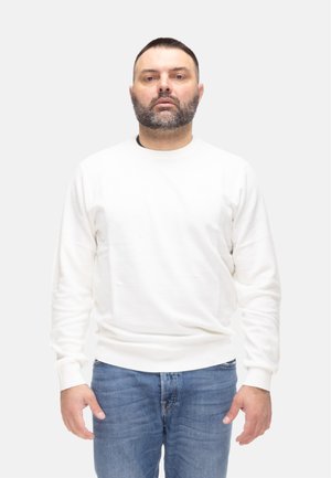 Uomo con capelli corti e barba, che indossa una camicia bianca a maniche lunghe e jeans blu, in piedi davanti a uno sfondo chiaro semplice.