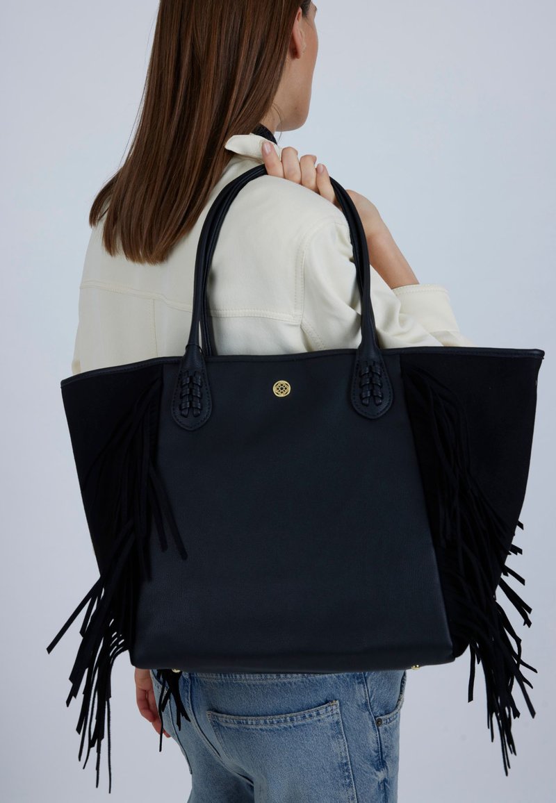 Musta olkalaukku hapsuilla, jossa on sileä nahkamateriaali ja punotut yksityiskohdat kahvoissa. Yksinkertainen kultainen pyöreä logo näkyvissä.