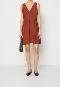 Robe sans manches couleur rouille avec un décolleté en V, des fronces sous la poitrine et une jupe fluide. Associée à un sac à main noir et des chaussures noires.