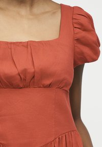 Κοντινό πλάνο ενός ατόμου που φοράει φόρεμα σε σκουρό πορτοκαλί απόχρωση με τετράγωνο λαιμόκοψη και φουσκωτά κοντά μανίκια, εμφανίζοντας την υφή του υφάσματος και τις πιέτες.