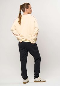 Žena s copom, oblečená v krémovej mikine, čiernych teplákoch a bielych teniskách, stojaca bokom s rukami za chrbtom.