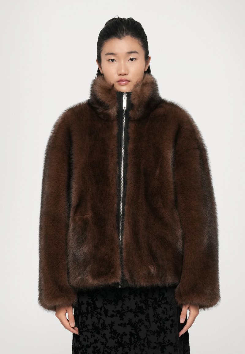 Veste en fausse fourrure marron avec un col montant, fermeture éclair sur le devant et poches latérales. Texture douce et coupe décontractée, sur un fond uni.