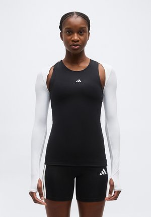 Czarnoskóra sportsmenka ubrana w czarny top i spodenki Adidas oraz białe opaski kompresyjne, stojąca na jednolitym tle.