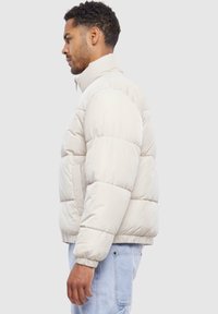 Jachetă puffer în bej deschis cu guler înalt, design matlasat, manșete elastice și buzunare laterale. Textură moale și izolație pentru căldură.