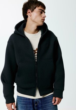 Schwarze Zip-Hoodie aus Baumwollmischung, mit Kängurutasche, gerippten Bündchen und weichem Griff, getragen über einem cremfarbenen Pullover.