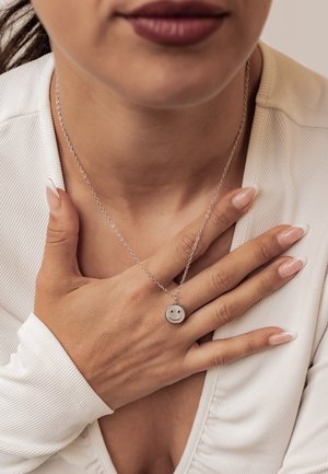 Woman with dark lipstick wearing a white top, hand on chest, showing long nails and a silver necklace with a smiley face pendant.