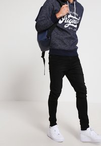 Felpa con cappuccio blu navy con scritte bianche, jeans neri, sneakers bianche e zaino blu. Design aderente con coulisse e polsini a costine.