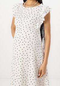 Nainen pukeutunut valkoiseen äitiysmekkoon, jossa on mustia pilkkuja ja röyhelöhihoja, asusteena kerrokselliset helmistä ja kullasta tehdyt kaulaketjut.