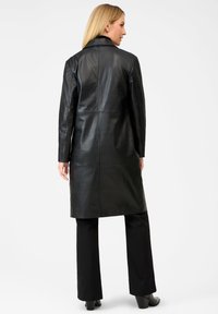Vrouw met blond haar, gekleed in een lange zwarte leren jas, zwarte broek en zwarte hoge hakken, staande tegen een witte achtergrond.