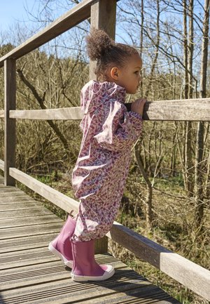 Lichtroze regenjas met bloemenpatroon, lange mouwen en een capuchon, gecombineerd met roze regenlaarzen met witte accenten, staand op een houten loopbrug.