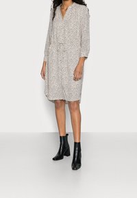 Langärmliges, locker sitzendes Kleid mit V-Ausschnitt, welches einen cremefarbenen Hintergrund und schwarze Punkte hat. Kombiniert mit schwarzen Stiefeletten.