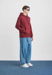 Felpa bordeaux con tasca, vestibilità comoda. Jeans a gamba larga azzurri. Sneakers bianche. Gli accessori includono occhiali da sole blu. Design minimalista.