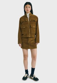 Braune Cordjacke mit zwei vorderen Taschen, kombiniert mit einem passenden Rock. Das Model trägt einen weißen Rollkragenpullover und schwarze Sneakers mit weißen Schnürsenkeln.