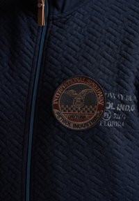 Jachetă bleumarin cu captuseală, cu fermoar, dintr-un material texturat, având un patch brodat circular cu un design de vultur și text, accentuată de un fermoar albastru.