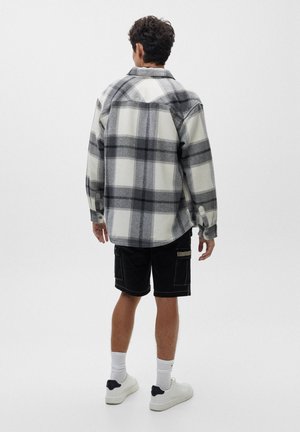 Mann trägt übergroßes graues und weißes kariertes Hemd, schwarze Cargoshorts, weiße Socken und weiße Sneakers, steht mit dem Rücken zur Kamera vor einfarbigem Hintergrund.