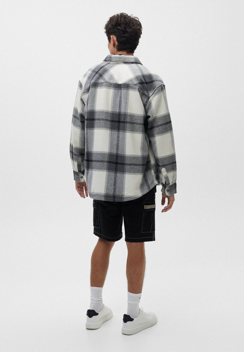 Homme portant une chemise à carreaux grise et blanche oversize, un short cargo noir, des chaussettes blanches et des baskets blanches, debout de dos sur un fond uni.