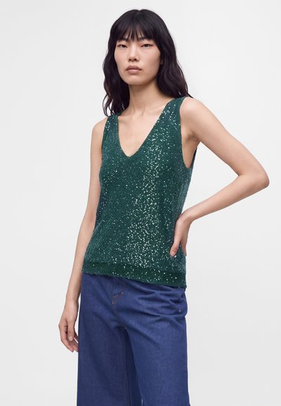 Jeune femme aux longs cheveux foncés portant un haut vert à sequins sans manches et un jean bleu décontracté, debout avec une main sur la hanche.
