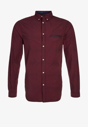 Uomo che cammina, indossa una camicia bordeaux con bottoni, jeans azzurri chiari, scarpe nere e tiene uno zaino nero per una spallina.
