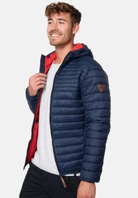Navy blue puffer jacket with red interior, quilted design, hood, and a leather logo patch on the left shoulder. Zipper front closure.