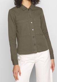 Olivově zelená košile s knoflíky vyrobená z bavlny, s límečkem, dvěma náprsními kapsami a dlouhými rukávy, v kombinaci s bílými kalhotami.