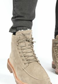 Beige mockaskängor med en texturerad yta, med bruna gummisulor, snörning och subtil branding på tungan.