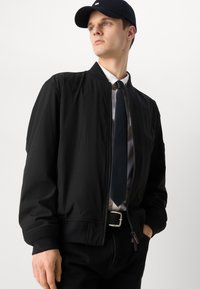 Černá bomber bunda se zipem, s žebrovanými manžetami a lemem, nošena přes bílou košili a kravatu. V doprovodu černé čepice.