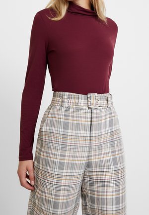 Femme portant un haut bordeaux ajusté à manches longues, rentré dans un pantalon taille haute à carreaux multicolores avec ceinture, sur fond blanc.