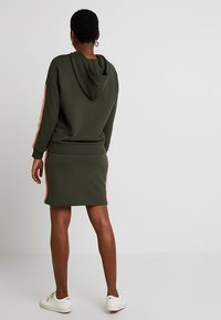 Un hoodie vert olive avec capuche et une jupe assortie, présentant des bandes latérales roses. Tissu lisse avec un design ajusté et une taille élastiquée.