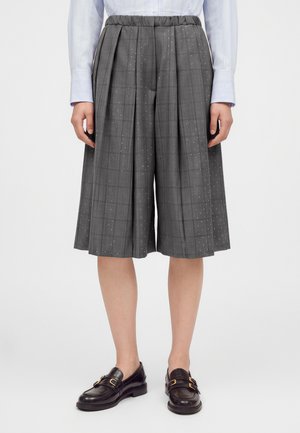 Personne portant un short plissé gris à jambes larges avec de petits pois blancs, une chemise bleu clair à manches longues et des mocassins noirs.