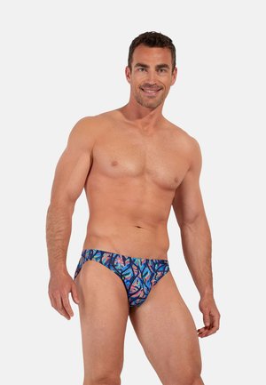 Glimlachende gespierde man met kleurrijke, met patronen bedrukte zwembroek, staand tegen een effen lichtgrijze achtergrond.