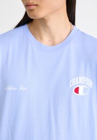 Gaiši zila kokvilnas T-kreklu ar apaļu kaklu, uz kura ir balta izšuvuma uzraksts "Athletics Dept." un "CHAMPION" logotips ar sarkanām akcentiem.