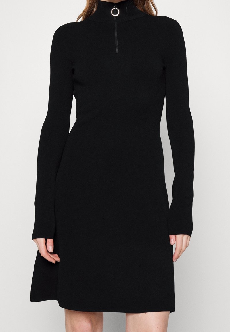 Langärmeliges schwarzes Strickkleid mit hohem Kragen und silbernem Reißverschlussdetails am Hals, getragen von einer Person, die vor einem weißen Hintergrund steht.