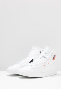 Valkoiset korkeavartiset tennarit, joissa on teksturoitu kangaspäällinen, oranssi Nike-logo, mustat sivu-akseptit ja mustat nauhat; kumipohja.