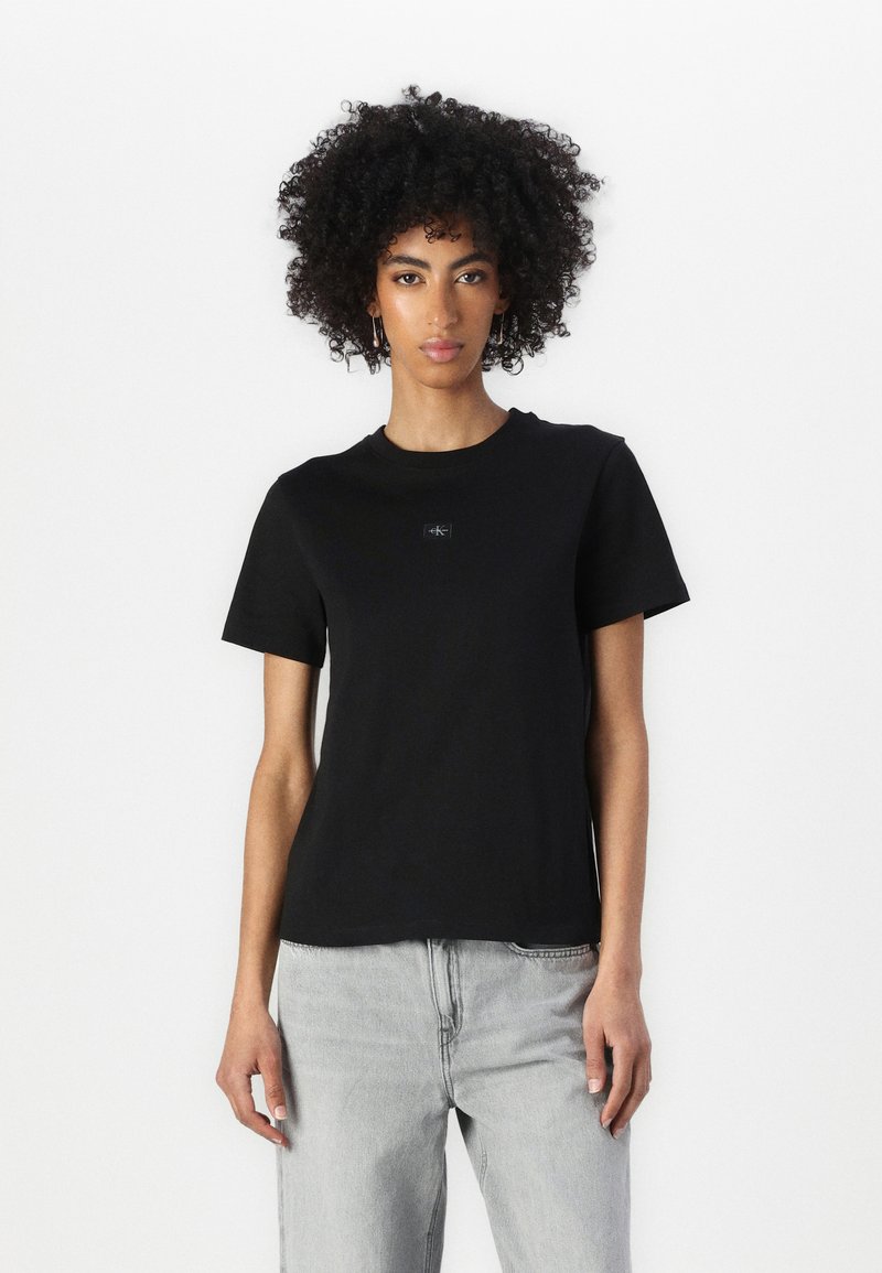 Svart bomull t-skjorte med korte ermer og rund hals, med en liten logo på brystet. Brukt sammen med lysegrå jeans.
