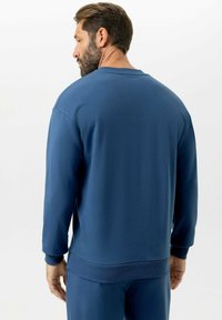 Bärtiger Mann mit braunen Haaren, der einen blauen Langarm-Sweatshirt und passende blaue Hose trägt, mit dem Rücken zum Betrachter vor einem weißen Hintergrund.