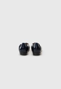Pantofi din piele lacuită de culoare bleumarin, cu vârf rotund, detaliu decorativ sub formă de fundiță și toc mic negru, văzuți din spate.