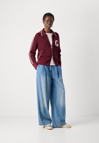 Giacca zip-up a coste color bordeaux con strisce bianche e logo 'G', abbinata a jeans denim azzurri svasati. Scarpe argento e beige.