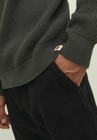 Dunkelgrauer, gerippter Pullover mit einem weißen und roten Logodetail am Saum, kombiniert mit schwarzen Hosen. Strukturiertes Material; taillierte Passform.