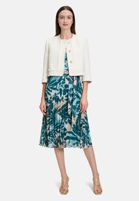 Biała kurtka z tweedu, krótka, z złotymi guzikami, noszona na turkusowej sukience w kwiatowy wzór, z plisami i zawiązanym w pasie, uzupełniona żółtymi sandałami.