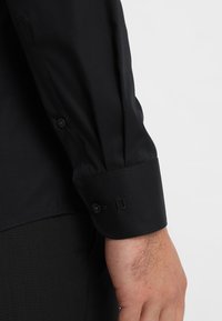 Svart långärmad skjorta med strukturerad krage, knäppt ärmslut, slät textur och skräddarsydd passform, visad vid ärmen och handleden.