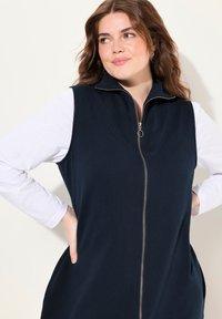 Gilet senza maniche di colore blu navy con colletto alto e zip frontale, caratterizzato da una texture di tessuto liscia e tasche laterali per una maggiore funzionalità.